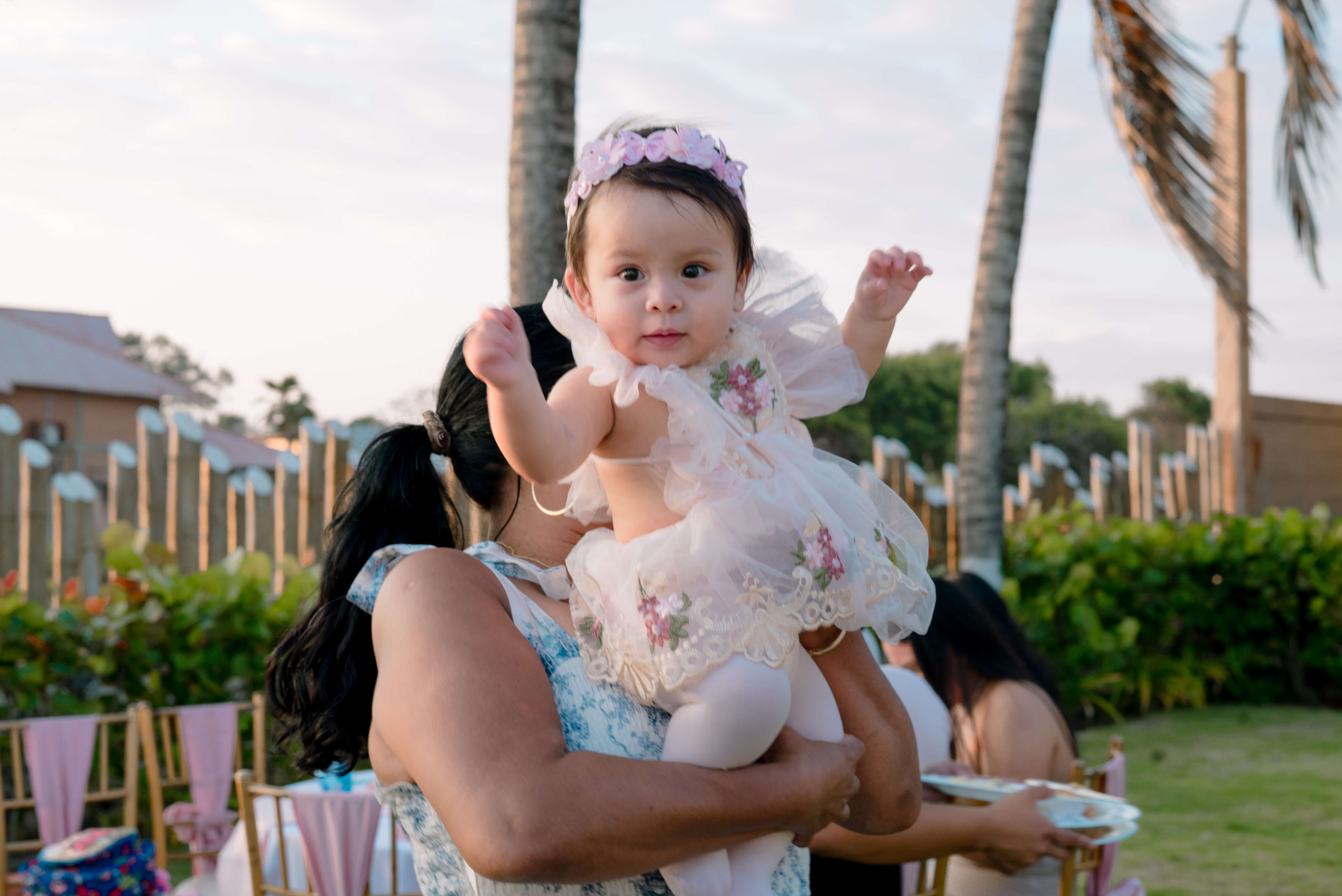 Bebé Mía de un año en brazos de su mamá con vestido de encaje rosado y diadema de flores, celebración al aire libre en Las Marías Playas al atardecer